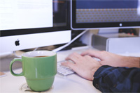 Escritorio con una taza de café, pantallas y manos sobre un teclado